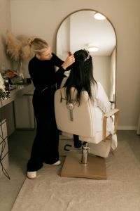 Welcome Home 105 A hairstylist adorns a bride's hair with floral accessories in a chic salon setting.